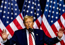 Man speaking on stage with American flags behind