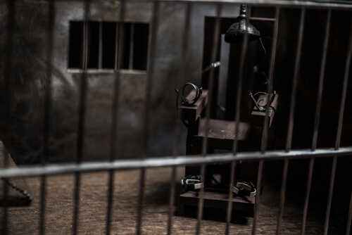 An electric chair inside a dimly lit prison cell with bars in the foreground