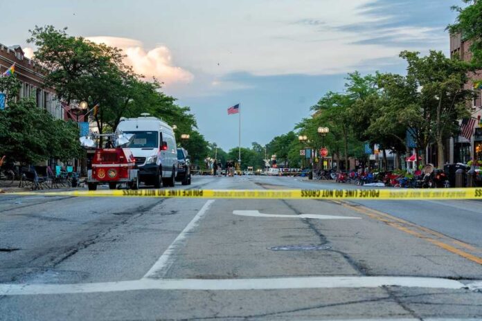 Police tape blocking a street with emergency vehicles and an American flag in the background