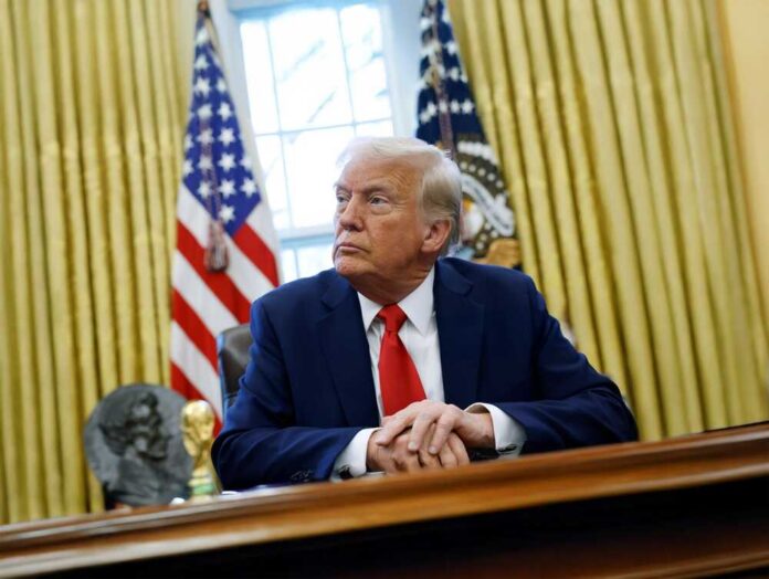 shutterstock_2644275941.jpg A man in a suit sitting at a desk in the Oval Office with American flags in the background