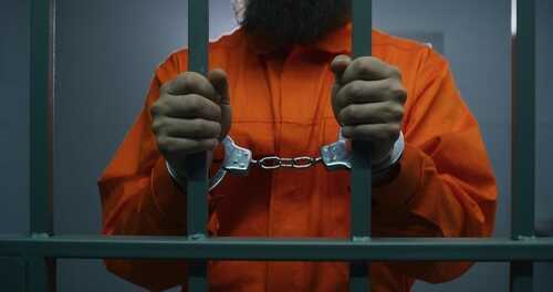 A prisoner in an orange jumpsuit holding jail bars with handcuffs