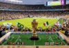 World Cup SECURITY CRISIS — Total DHS Collapse A miniature soccer field with a World Cup trophy in front of a cheering crowd